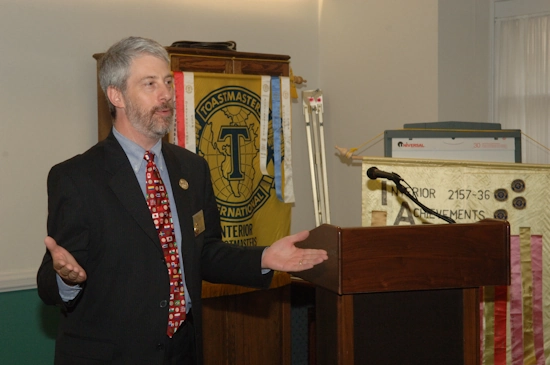 A man giving a speech next to a podium. Behind him is a banner with the Toastmasters International logo.