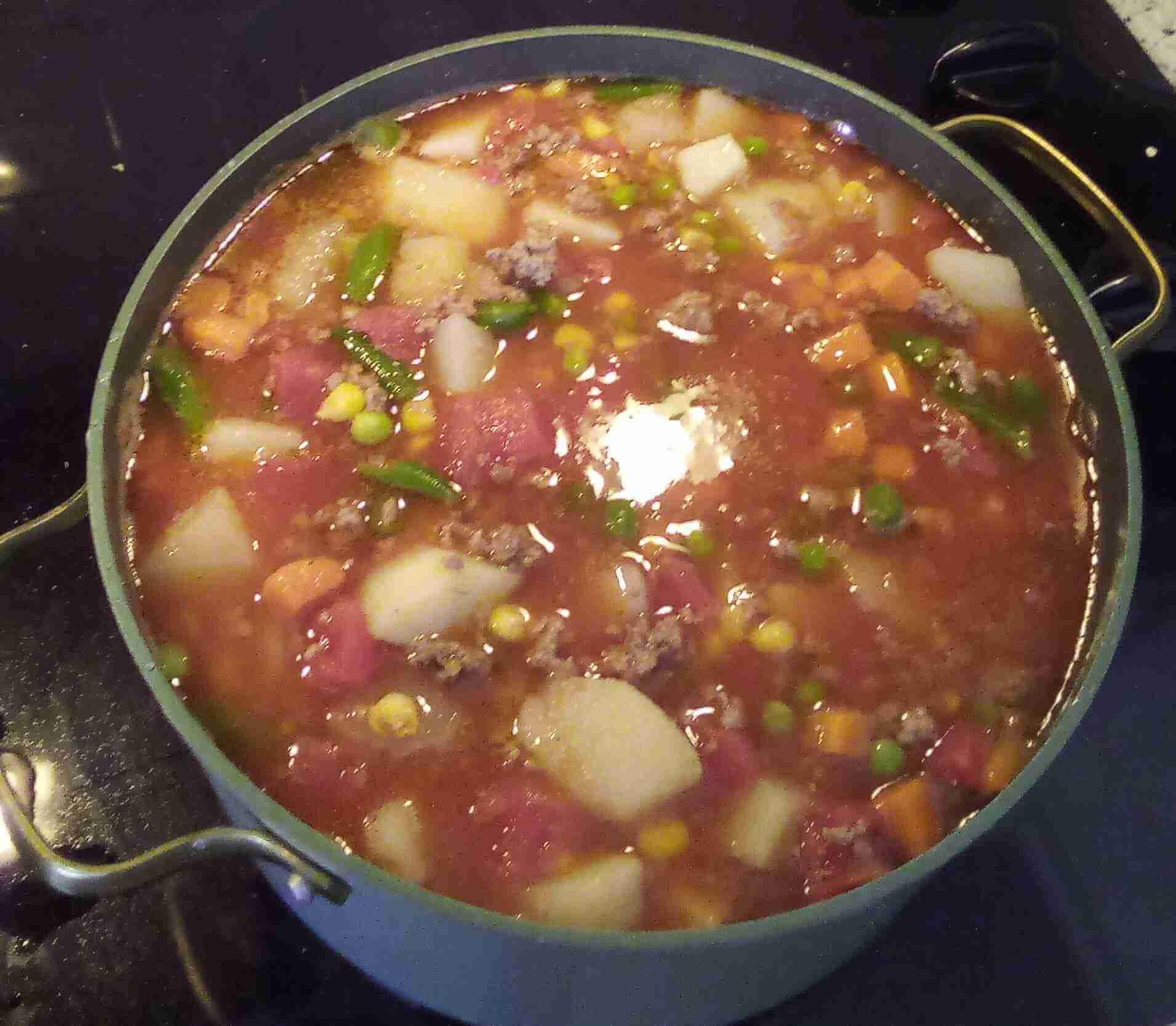 A teal pot sitting on a black glass stove. The pot is filled with a tomato base vegetable soup with course cut potatoes, carrots, and smaller chunks of other vegetables alongside cooked ground beef