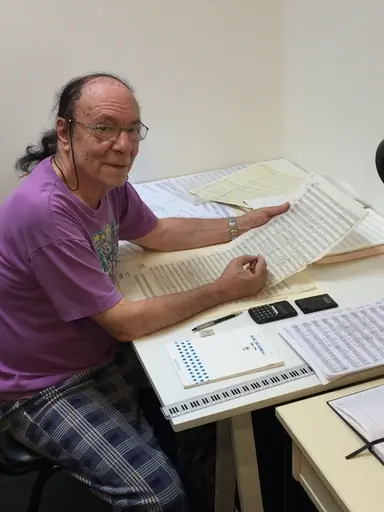 A photo of Jorge Antunes composing on a large orchestral score at a wide desk.