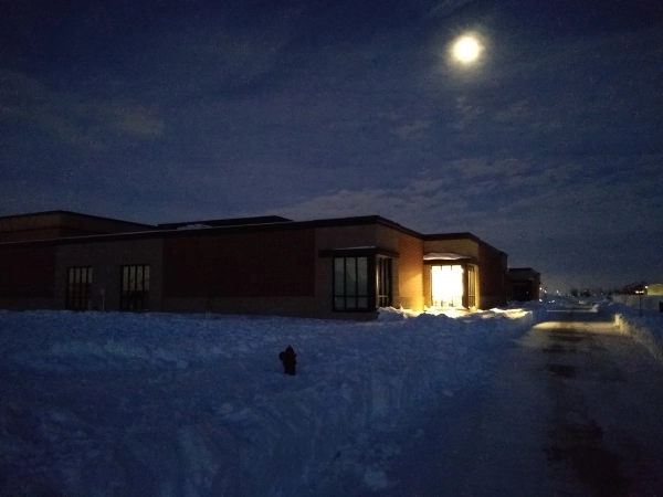 A school with one room lit up, surrounded by snow. The full moon is visible behind clouds