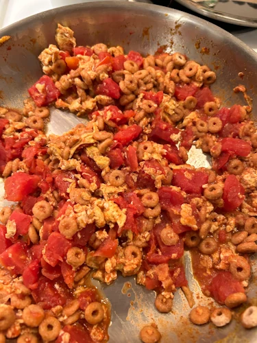 A pan containing a mixture of scrambled eggs, diced tomatoes, and cheerios.