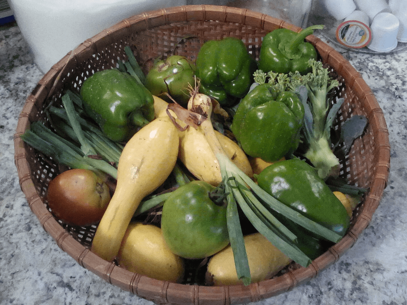 A brown woven basked filled with bell peppers, green peas, squache, onions, and several tomato in varying states of ripeness